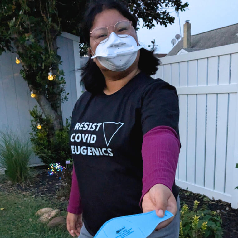 Model wearing a black tshirt. The t-shirt has white text across the chest "RESIST COVID EUGENICS" with each word stacked on top of each other in left alignment. To the right of the words is a white outline of an upside-down black triangle.